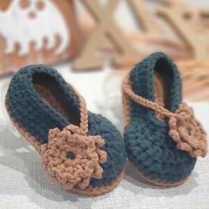Pu&ograve; includere: Un paio di scarpine per neonati all'uncinetto in blu scuro e marrone. Le scarpine hanno un dettaglio floreale sul davanti e un cinturino sulla parte superiore.