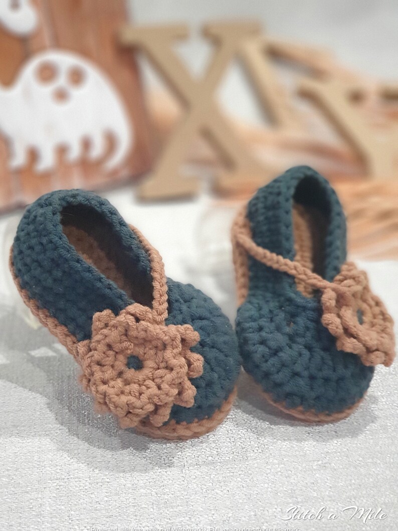 Pu&ograve; includere: Un paio di scarpine per neonati all'uncinetto in blu scuro e marrone. Le scarpine hanno un dettaglio floreale sul davanti e un cinturino sulla parte superiore.