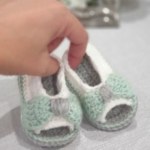 Pode incluir: Um par de sapatos de beb&eacute; de croch&ecirc; em verde, branco e cinza. Os sapatos t&ecirc;m um design de ponta aberta e um la&ccedil;o na frente.
