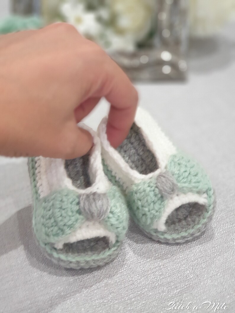 Pode incluir: Um par de sapatos de beb&eacute; de croch&ecirc; em verde, branco e cinza. Os sapatos t&ecirc;m um design de ponta aberta e um la&ccedil;o na frente.