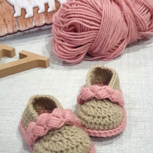 Pu&ograve; includere: Un paio di scarpine per neonati all'uncinetto in beige con rifiniture rosa. Le scarpine hanno un cinturino rosa intrecciato sulla parte superiore. Un gomitolo di filato rosa &egrave; sullo sfondo.