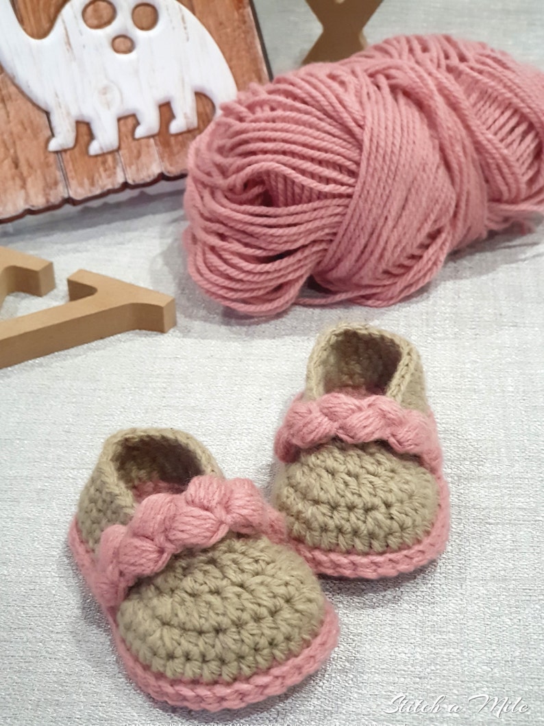 Pu&ograve; includere: Un paio di scarpine per neonati all'uncinetto in beige con rifiniture rosa. Le scarpine hanno un cinturino rosa intrecciato sulla parte superiore. Un gomitolo di filato rosa &egrave; sullo sfondo.