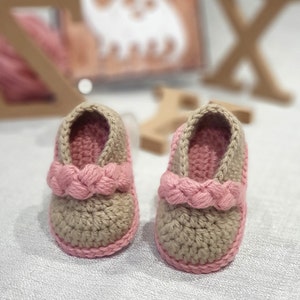 Pu&ograve; includere: Un paio di scarpine per neonati all'uncinetto in beige e rosa. Le scarpine hanno un cinturino rosa intrecciato sulla parte superiore.