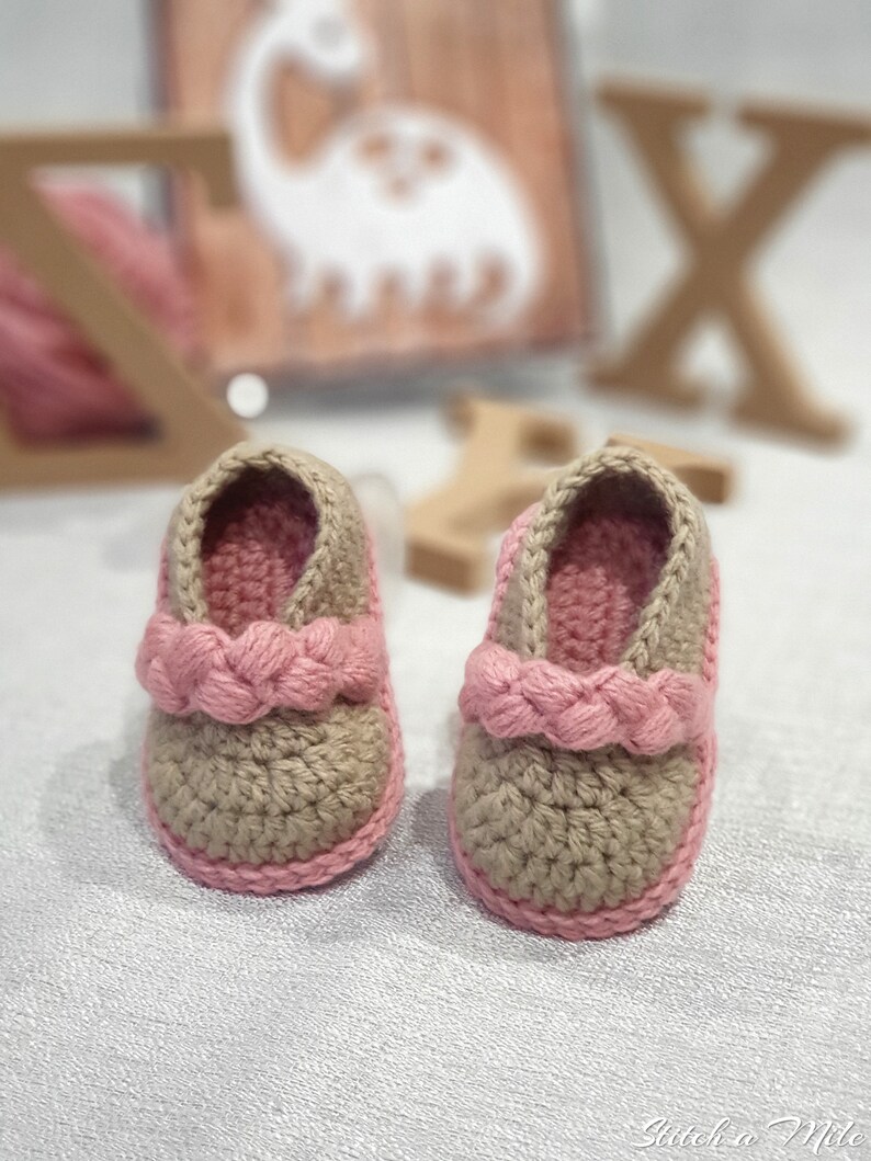 Pu&ograve; includere: Un paio di scarpine per neonati all'uncinetto in beige e rosa. Le scarpine hanno un cinturino rosa intrecciato sulla parte superiore.