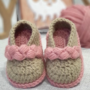 Pu&ograve; includere: Un paio di scarpine per neonati all'uncinetto in beige e rosa. Le scarpine hanno un cinturino rosa intrecciato sulla parte superiore e un bordo rosa attorno alla parte inferiore.