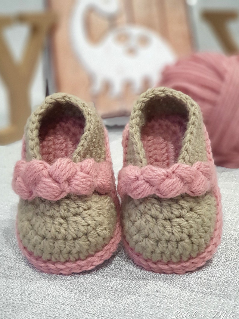 Pu&ograve; includere: Un paio di scarpine per neonati all'uncinetto in beige e rosa. Le scarpine hanno un cinturino rosa intrecciato sulla parte superiore e un bordo rosa attorno alla parte inferiore.