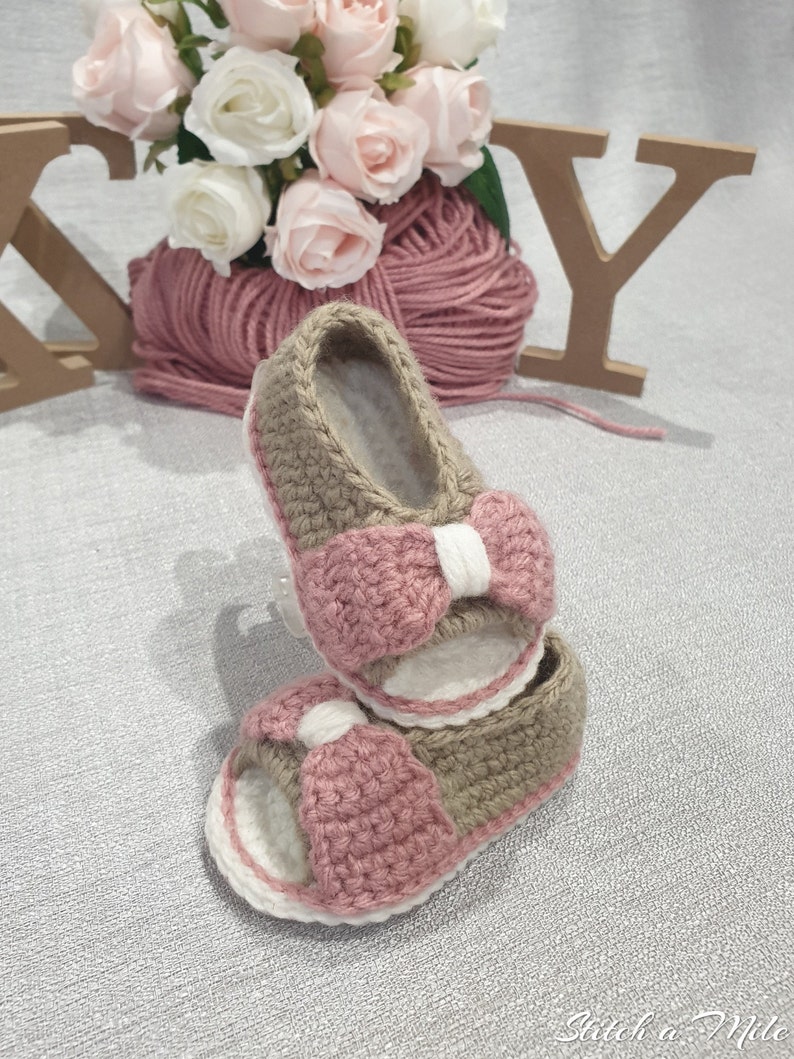 Pode incluir: Um par de sand&aacute;lias de beb&eacute; de croch&ecirc; com la&ccedil;os cor-de-rosa e um esquema de cores castanho e branco.