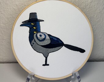 May include: Embroidered artwork of a magpie wearing a black hat, with three yellow eyes. The bird is depicted in shades of blue, gray, and black, set against a white background within a wooden embroidery hoop. The artwork is displayed on a clear plastic stand.