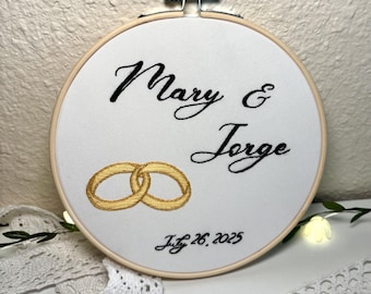 May include: White embroidery hoop with the text "Mary & Jorge" in black cursive script and two gold rings intertwined. The date "July 26, 2025" is written below the rings.