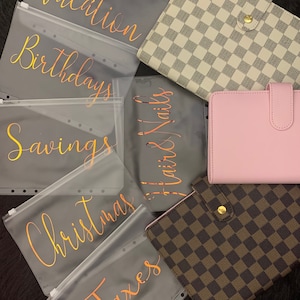 May include: Six clear plastic zippered pouches with gold lettering. The pouches are labeled "Vacation", "Birthdays", "Savings", "Christmas", "Taxes", and "Hair & Nails".  There are three different patterned binders in the background. One is white with a black checkered pattern, one is pink, and one is brown with a tan checkered pattern.