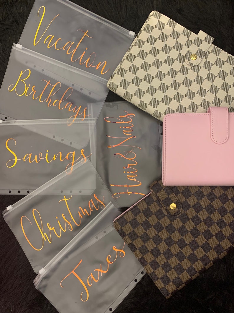 May include: Six clear plastic zippered pouches with gold lettering. The pouches are labeled "Vacation", "Birthdays", "Savings", "Christmas", "Taxes", and "Hair & Nails".  There are three different patterned binders in the background. One is white with a black checkered pattern, one is pink, and one is brown with a tan checkered pattern.