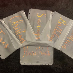 May include: Six clear plastic zippered pouches with gold lettering. The pouches are labeled "Taxes", "Christmas", "Savings", "Birthday", "Vacation", and "Hair & Nails".