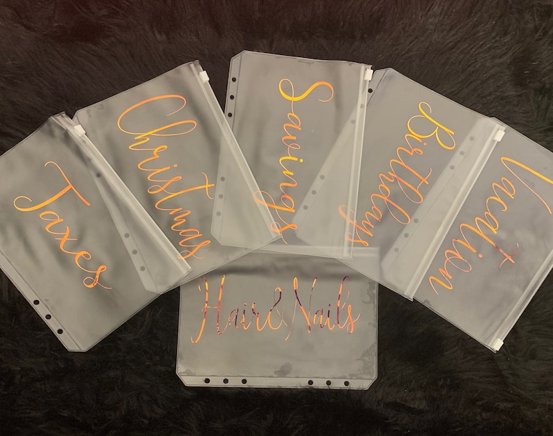 May include: Six clear plastic zippered pouches with gold lettering. The pouches are labeled "Taxes", "Christmas", "Savings", "Birthday", "Vacation", and "Hair & Nails".