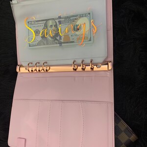 May include: A pink leather bound planner with a clear plastic pocket on the front. The pocket contains a $100 bill with the word "Savings" written in gold script on it.