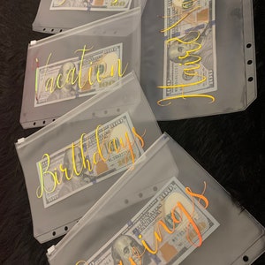 May include: Five clear plastic zippered pouches with gold lettering. Each pouch has a different label: "Taxes", "Vacation", "Birthday", "Savings", and "Home". Each pouch contains a US dollar bill.