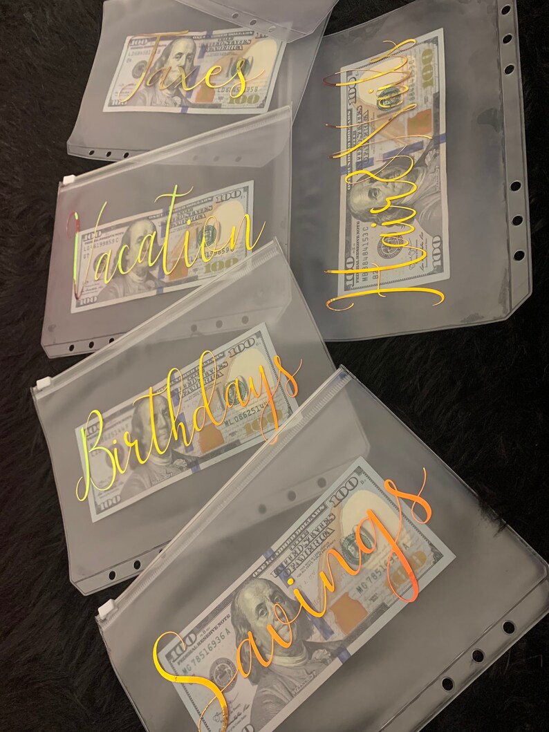 May include: Five clear plastic zippered pouches with gold lettering. Each pouch has a different label: "Taxes", "Vacation", "Birthday", "Savings", and "Home". Each pouch contains a US dollar bill.