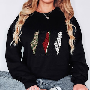 May include: Black sweatshirt featuring three stylized map outlines. One map is filled with a green and tan leaf pattern, another with a watermelon design, and the third with a black and white checkered pattern. The sweatshirt has a crew neck.