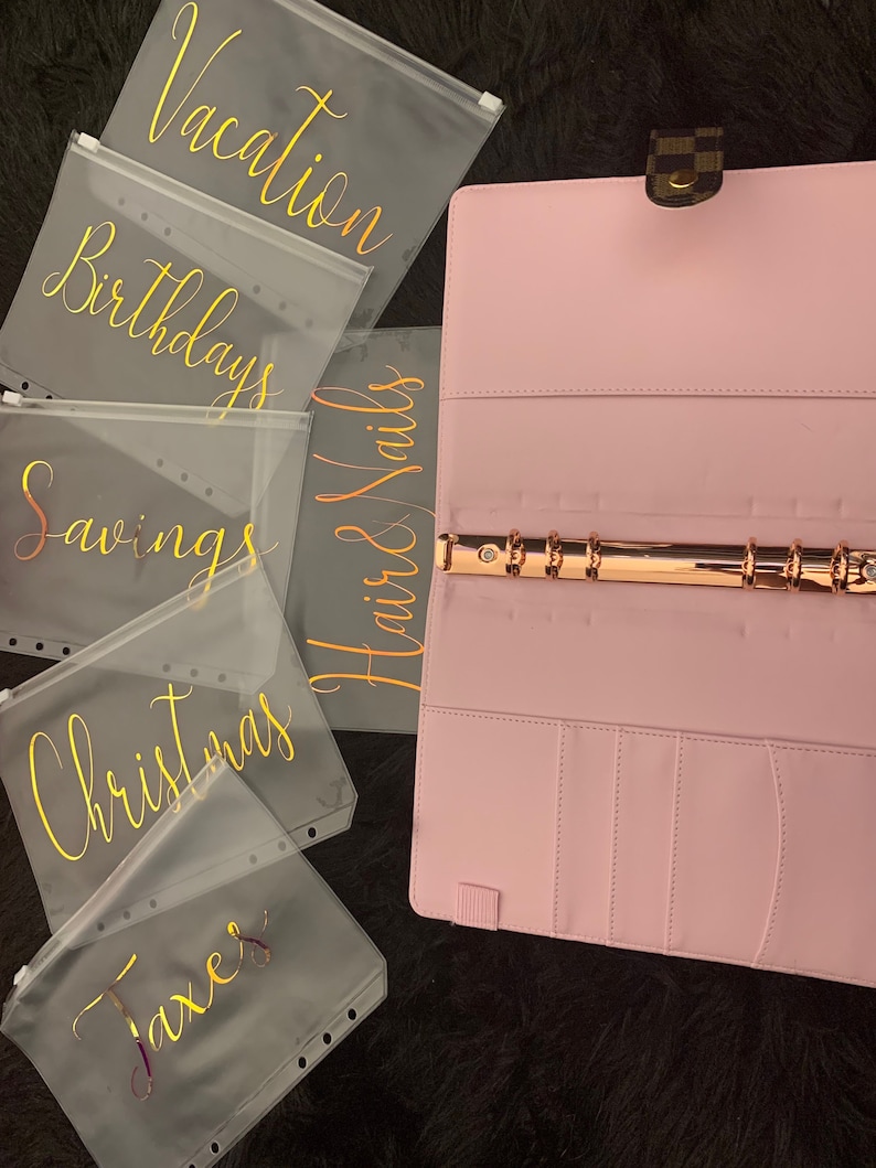 May include: Five clear plastic zippered pouches with gold lettering. The pouches are labeled "Vacation", "Birthdays", "Savings", "Christmas", and "Taxes". The pouches are sitting on a pink and brown background.