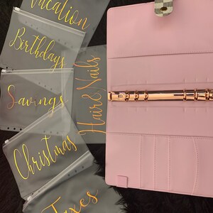 May include: Six clear plastic zippered pouches with gold lettering. The pouches are labeled "Vacation", "Birthdays", "Savings", "Hair & Nails", "Christmas", and "Taxes". The pouches are arranged on a black surface next to a pink binder with a gold ring.