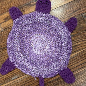 May include: A handmade crochet turtle in shades of purple and white. The turtle has a round body, four flippers, a head, and a tail. The crochet stitches create a textured surface. The turtle is a soft toy.