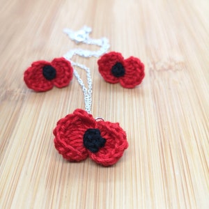 May include: A set of three crocheted red poppy flowers with black centres. The flowers are attached to a silver chain necklace and earrings.