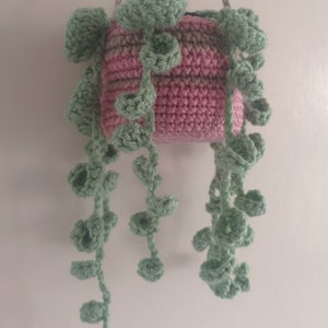 May include: A handmade crochet hanging planter in shades of pink and green. The planter is a square shape with a textured stitch, and the hanging vines are a light green colour. The planter is suspended by a beige rope.