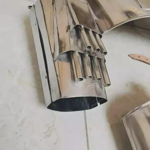 May include: A polished stainless steel exhaust tip featuring a large, wide-mouthed opening and a secondary internal section containing five smaller, cylindrical metal pipes arranged in a cluster. The metallic surface is highly reflective and smooth.