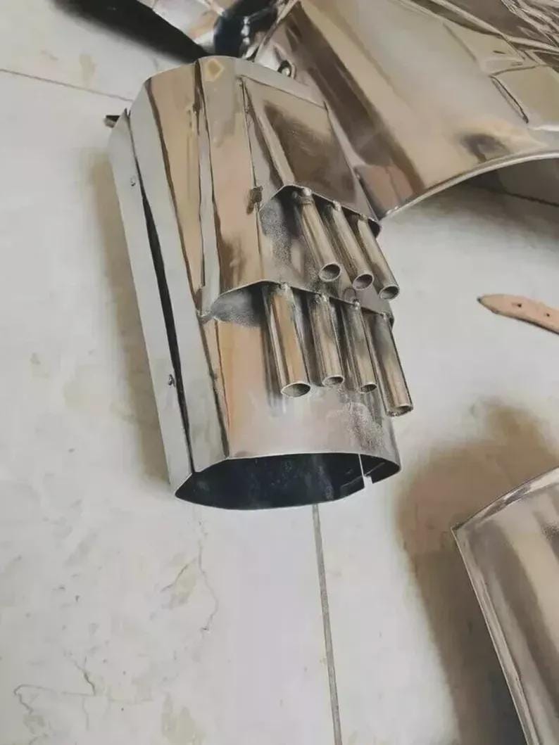 May include: A polished stainless steel exhaust tip featuring a large, wide-mouthed opening and a secondary internal section containing five smaller, cylindrical metal pipes arranged in a cluster. The metallic surface is highly reflective and smooth.