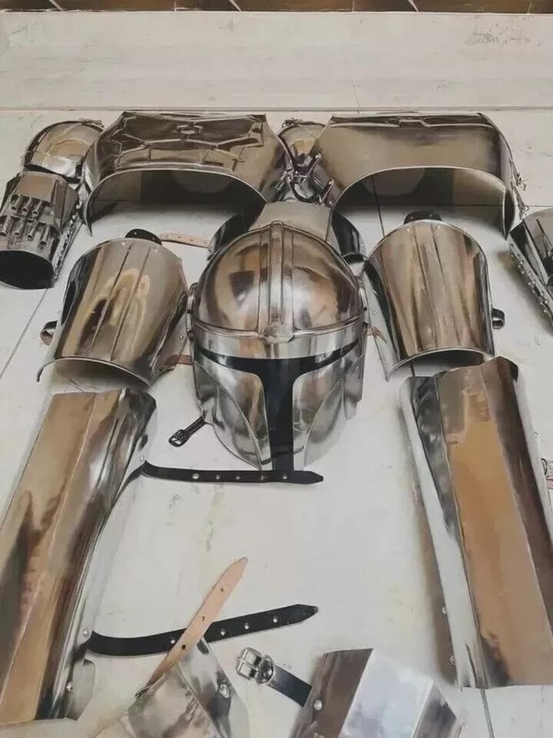 May include: A polished silver metal suit of armour set including a central helmet with a T-shaped visor, chest plates, shoulder guards, arm bracers, and leg greaves. The pieces feature black leather straps and metal buckles for fastening the components.