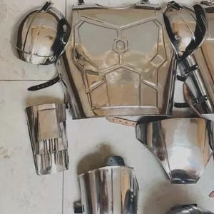 May include: A set of polished silver-coloured metal armour pieces including a chest plate with a central hexagonal emblem, two rounded shoulder pauldrons, arm guards, and leg greaves, all featuring black leather straps and metal buckles.