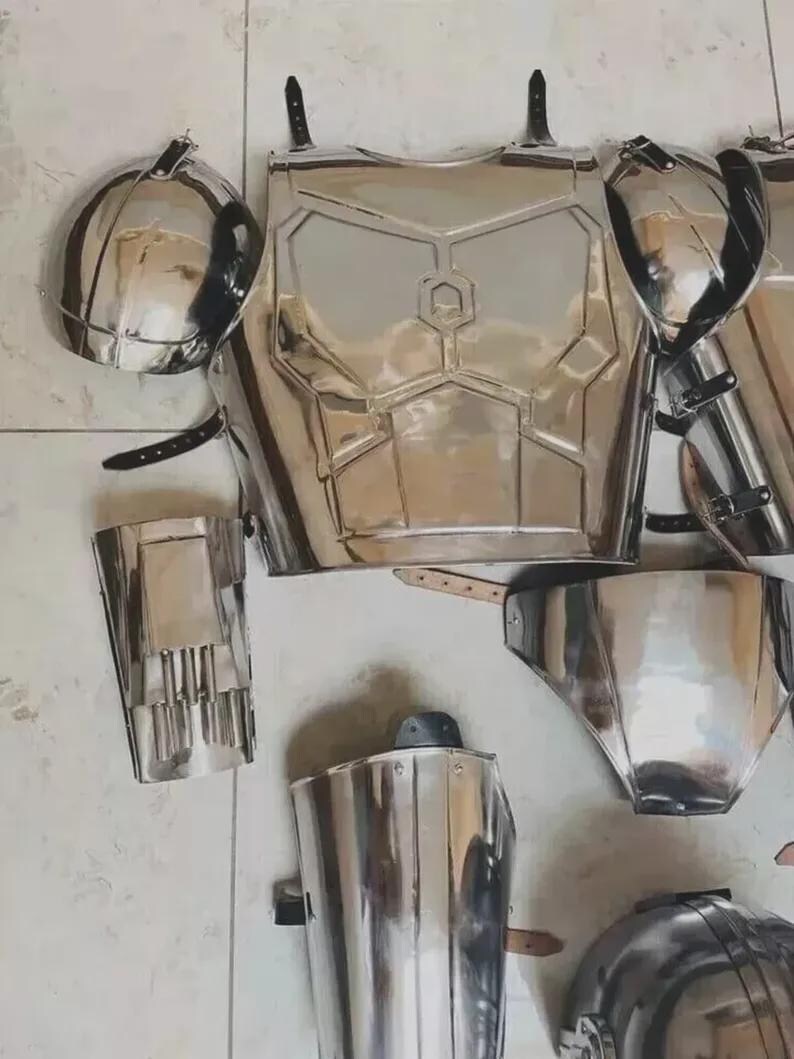 May include: A set of polished silver-coloured metal armour pieces including a chest plate with a central hexagonal emblem, two rounded shoulder pauldrons, arm guards, and leg greaves, all featuring black leather straps and metal buckles.