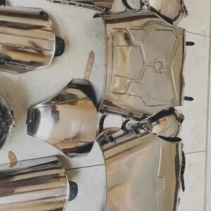 May include: A set of polished silver-coloured metal armour pieces including a chest plate, shoulder guards, arm bracers, and leg greaves. Each component features a reflective surface, structural ridges, and black leather straps with metal buckles.