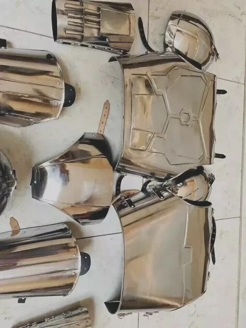 May include: A set of polished silver-coloured metal armour pieces including a chest plate, shoulder guards, arm bracers, and leg greaves. Each component features a reflective surface, structural ridges, and black leather straps with metal buckles.