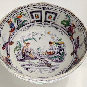 May include: A white ceramic bowl with a purple and green floral design. The bowl features a central scene with two figures in traditional clothing, a bird, and a vase. The rim of the bowl is decorated with a purple and black geometric pattern.
