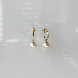Puede incluir: Un par de pendientes de aro dorados, cada uno con una perla redonda de color crema colgante. Los pendientes se muestran sobre una superficie blanca, con iluminación suave y sombras.