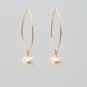 May include: A pair of gold-colored, curved drop earrings. Each earring features a delicate, elongated hook design with a small, round, cream-colored pearl pendant. The earrings are suspended against a plain, light background.