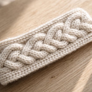 Può includere: Una fascia per capelli lavorata a maglia color crema con un motivo a treccia. La fascia è realizzata in un filato morbido e strutturato e misura circa 10 cm di larghezza. È esposta su una superficie di legno marrone chiaro.