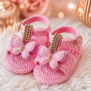 May include: A pair of pink crochet baby sandals. Each sandal features a sparkly butterfly applique and a strap with a gold buckle and jeweled accents. The sandals are displayed on a white, fluffy surface.