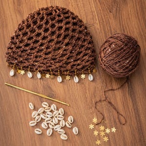 May include: A brown crocheted hat with a net-like design, adorned with seashells and gold accents. A ball of brown yarn, a crochet hook, seashells, and gold flower-shaped embellishments are arranged on a wooden surface, suggesting a craft project.