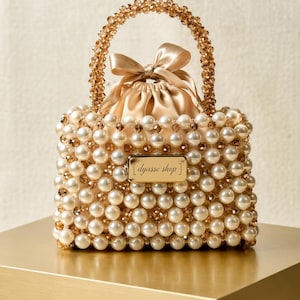 May include: A beaded handbag with a satin drawstring closure and a beaded handle. The bag is covered in cream-colored pearls and gold-colored beads. The handle is arched and the bag has a small rectangular tag that reads "dyasse shop".