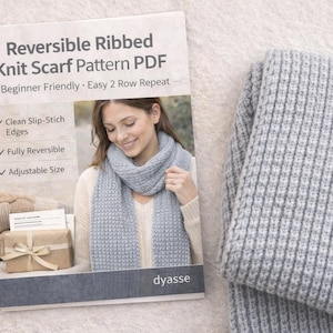 May include: A light gray, ribbed knit scarf and a pattern booklet titled "Reversible Ribbed Knit Scarf Pattern PDF" are displayed. The booklet highlights features like clean slip-stitch edges and adjustable size. Knitting needles and other accessories are visible.