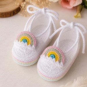 May include: A pair of white crocheted baby shoes featuring a rainbow applique on the front. The shoes are constructed with white yarn, have a rounded toe, and include white cord laces that tie at the top. The soles have subtle pastel stripes.