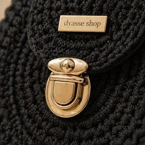 Op de afbeelding: Close-up van een zwarte gehaakte tas met een goudkleurige sluiting en een rechthoekig goudkleurig label met de tekst "dyasse shop". De tas heeft een getextureerde uitstraling.
