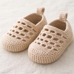 May include: A pair of beige crochet baby shoes. The shoes have a slip-on design with an open weave pattern on the top and a small loop at the heel. The shoes are made of a soft, textured yarn.