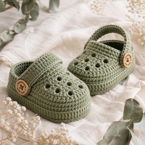 May include: A pair of olive green crochet baby shoes with a strap and wooden button detail. The shoes have a classic clog design with multiple holes. The wooden buttons are engraved with the words "handmade with love".