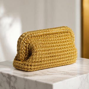 May include: A golden-yellow woven clutch bag with a rectangular shape and rounded edges. The bag is made of a textured material and sits on a white marble surface. The clutch has a simple, elegant design.