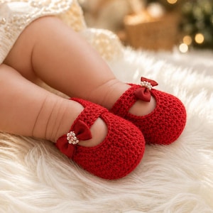 May include: A pair of bright red, crocheted baby shoes with a bow and pearl embellishment. The shoes have a Mary Jane style with a strap across the top. The shoes are on a fluffy, white surface.