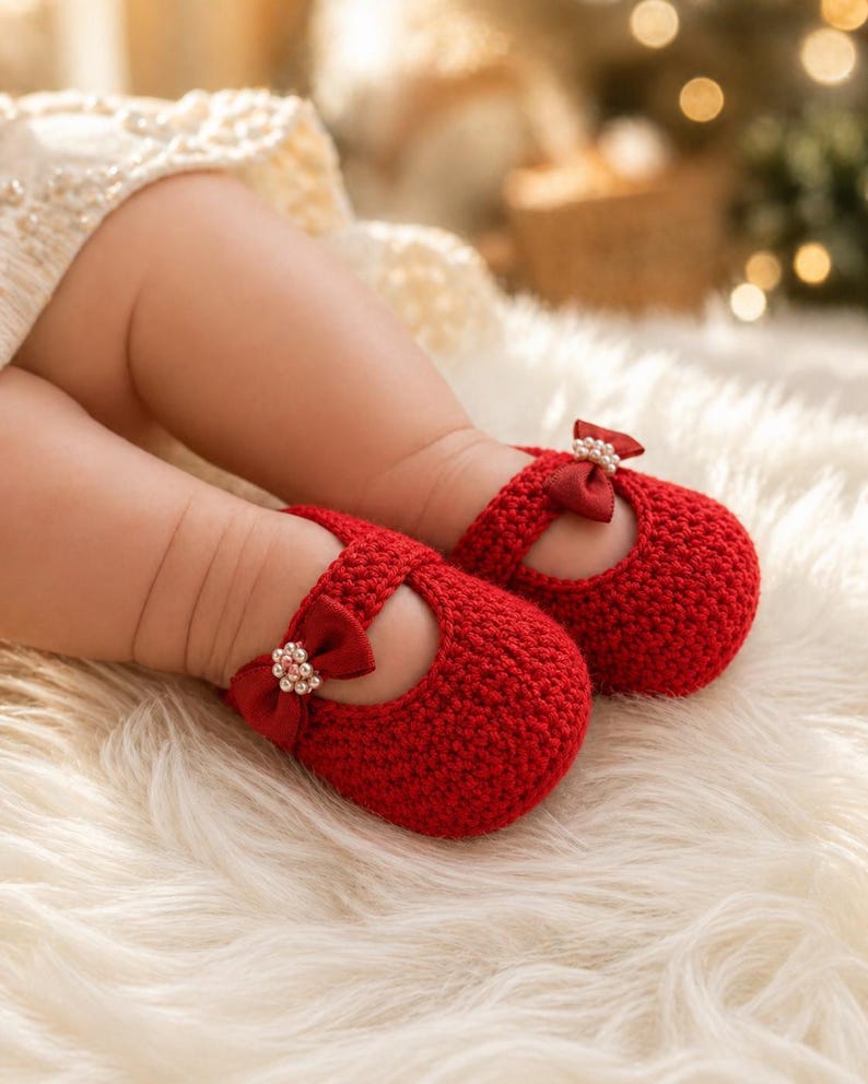 May include: A pair of bright red, crocheted baby shoes with a bow and pearl embellishment. The shoes have a Mary Jane style with a strap across the top. The shoes are on a fluffy, white surface.