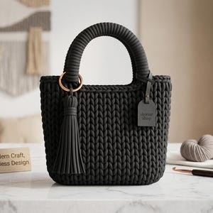 May include: A dark gray, hand-crocheted tote bag with a rounded handle and a decorative tassel. The bag features a copper-colored ring and a small tag that reads "dyasse shop". The bag is sitting on a white surface.