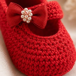 May include: A close-up of a red crocheted baby shoe. The shoe features a bow with a pearl embellishment. The shoe is a Mary Jane style, with a strap across the top of the foot. The crochet work is visible and detailed.
